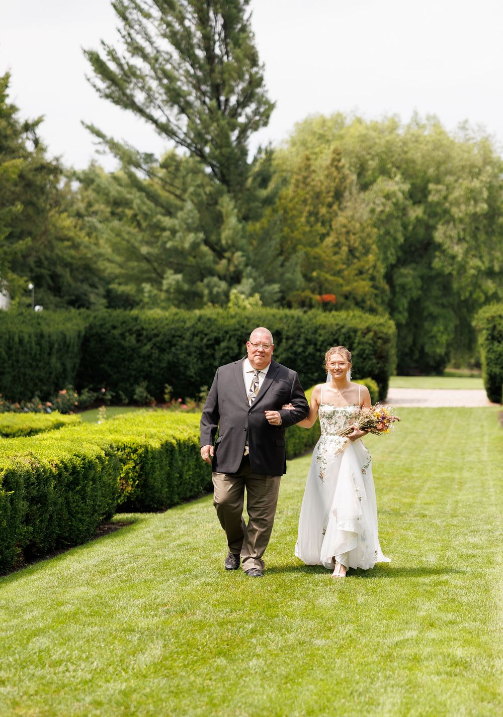 Bride in white gown walks with escort through lush green garden path on sunny wedding day.