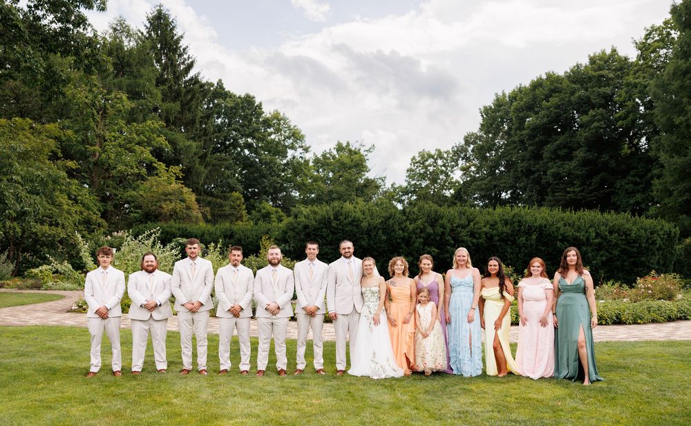 Wedding party poses outdoors in garden, groomsmen in white suits and bridesmaids in pastel gowns flanking bride and groom.