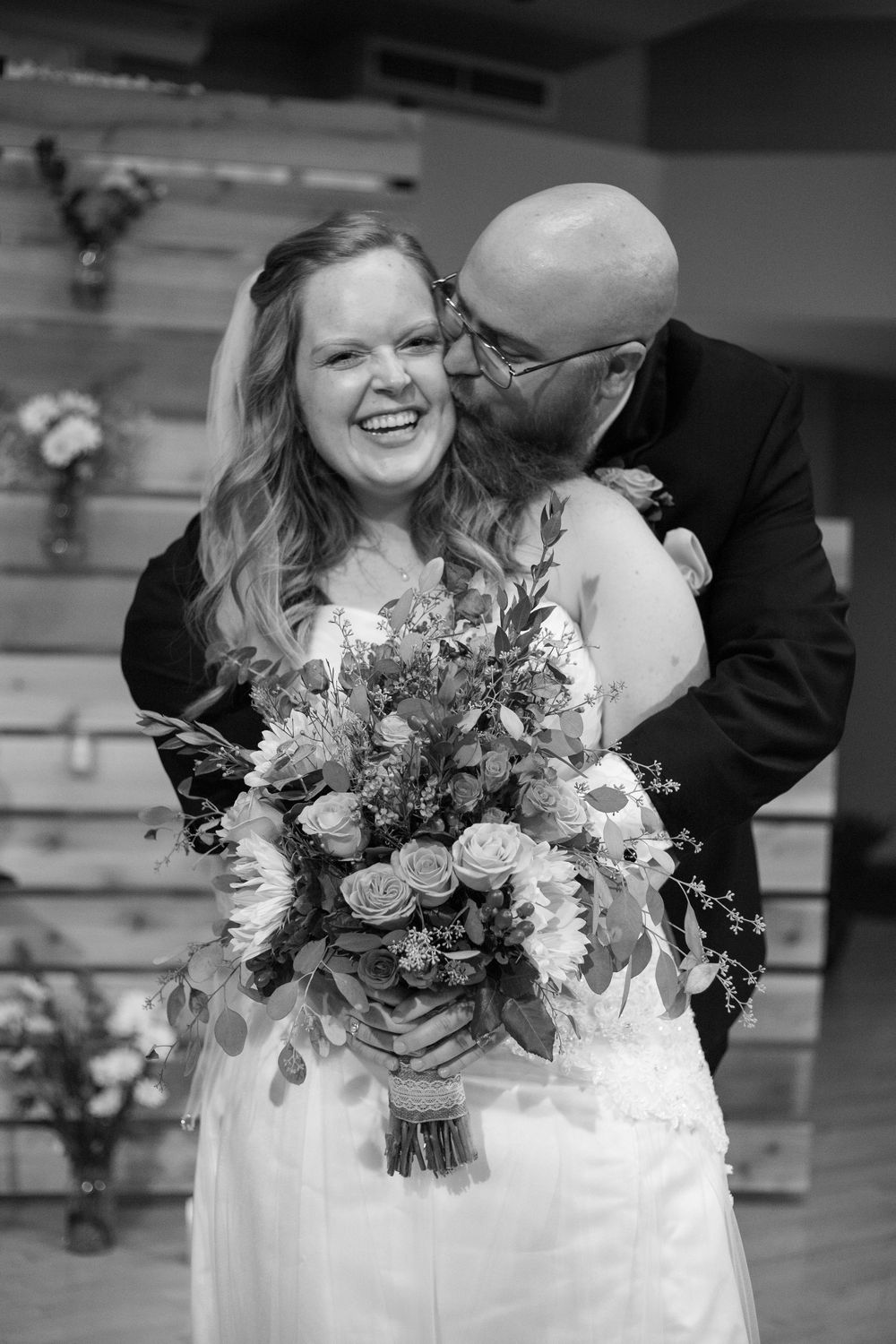 Groom kisses bride on cheek as she laughs holding floral bouquet in black and white wedding photo.