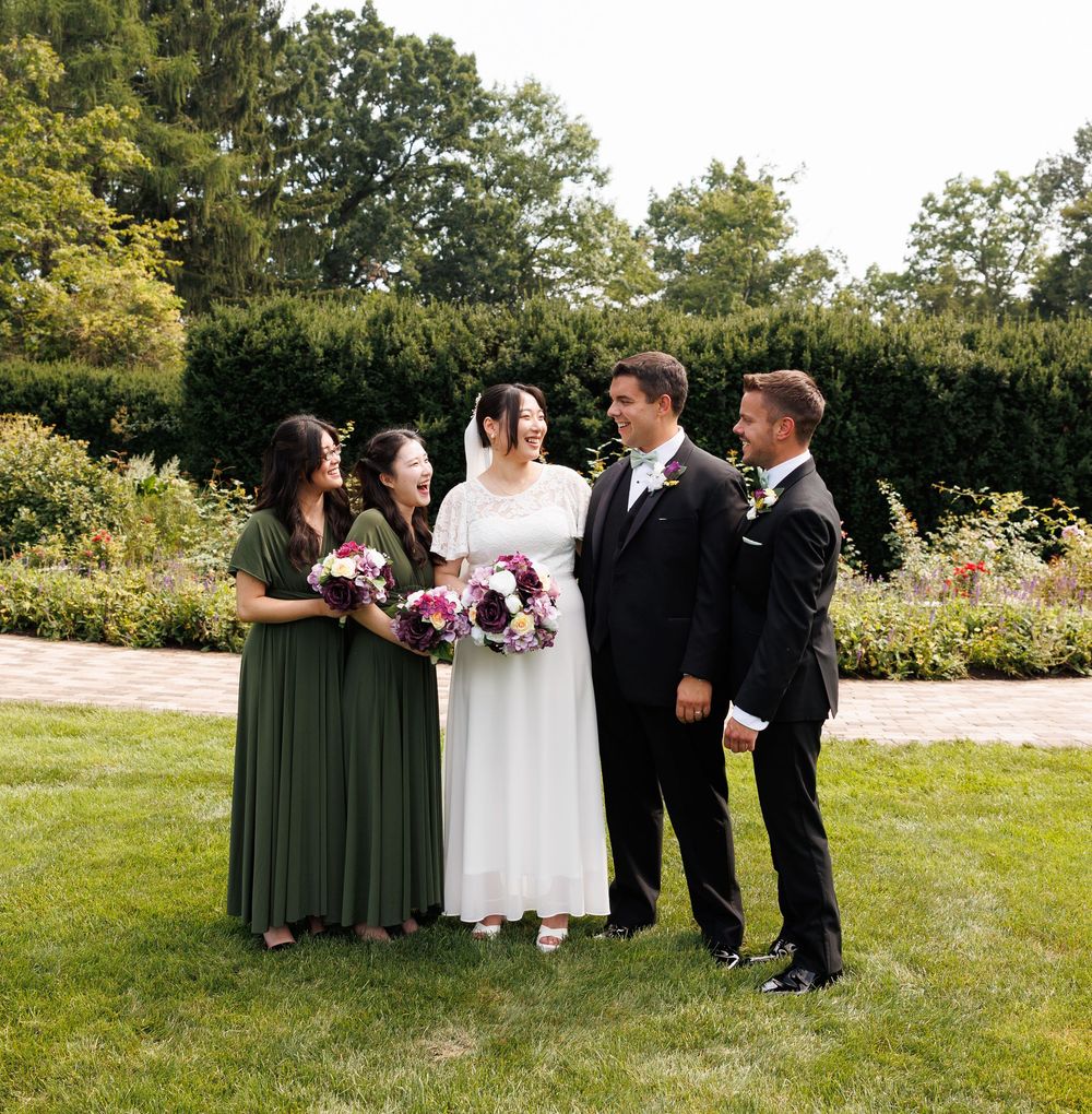 Wedding party of four poses outdoors on green lawn, bride in white gown holding bouquet, bridesmaids in olive green dresses.