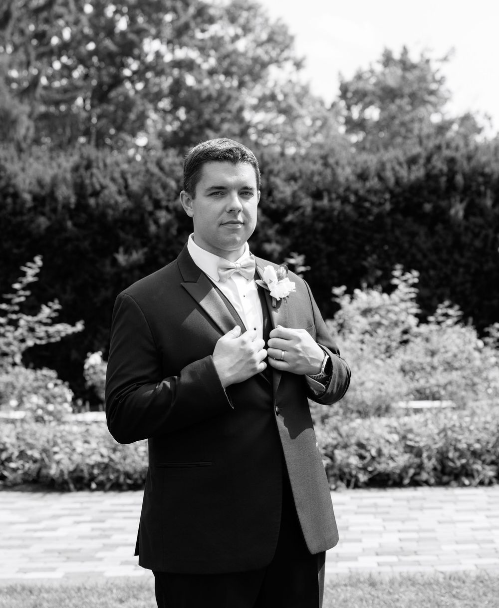 Black and white photo of a groom in a tuxedo adjusting his jacket outdoors with lush greenery background.