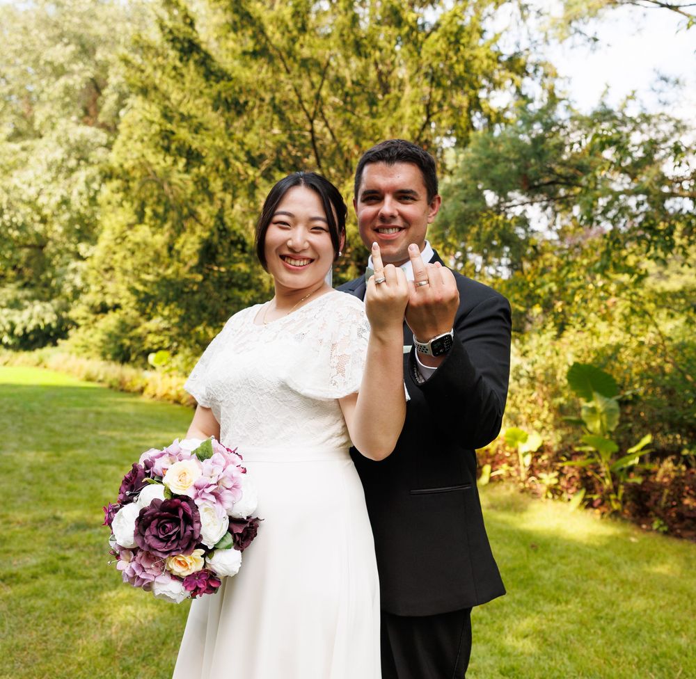 Newlywed couple smiling and showing wedding rings outdoors, bride holding purple and white bouquet in white lace dress.