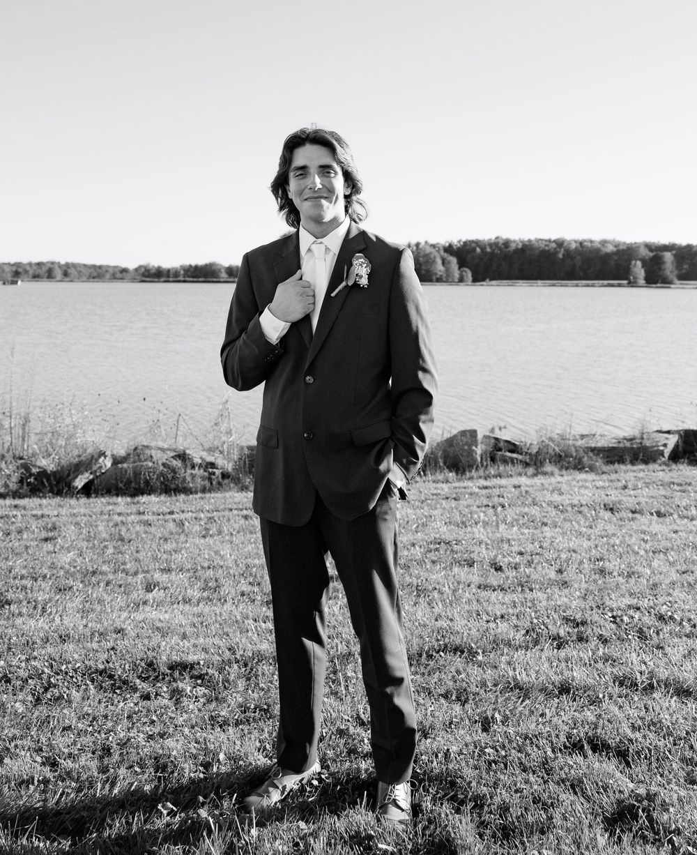 Black and white photo of a young man in a suit with boutonniere standing by a lake at an outdoor wedding.
