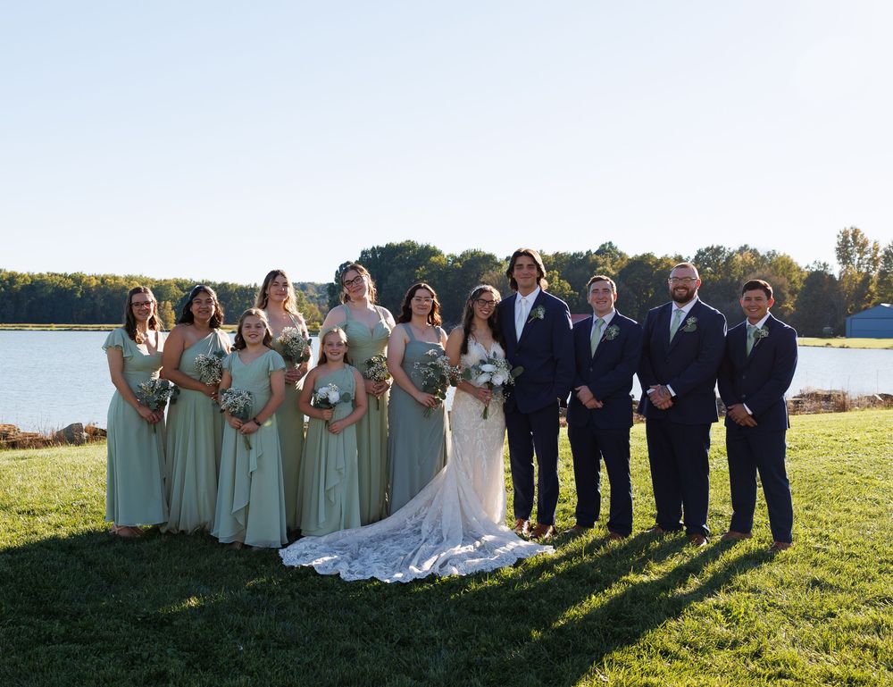 Wedding party poses outdoors by a lake, bridesmaids in sage green gowns and groomsmen in navy suits flanking the bride and groom.