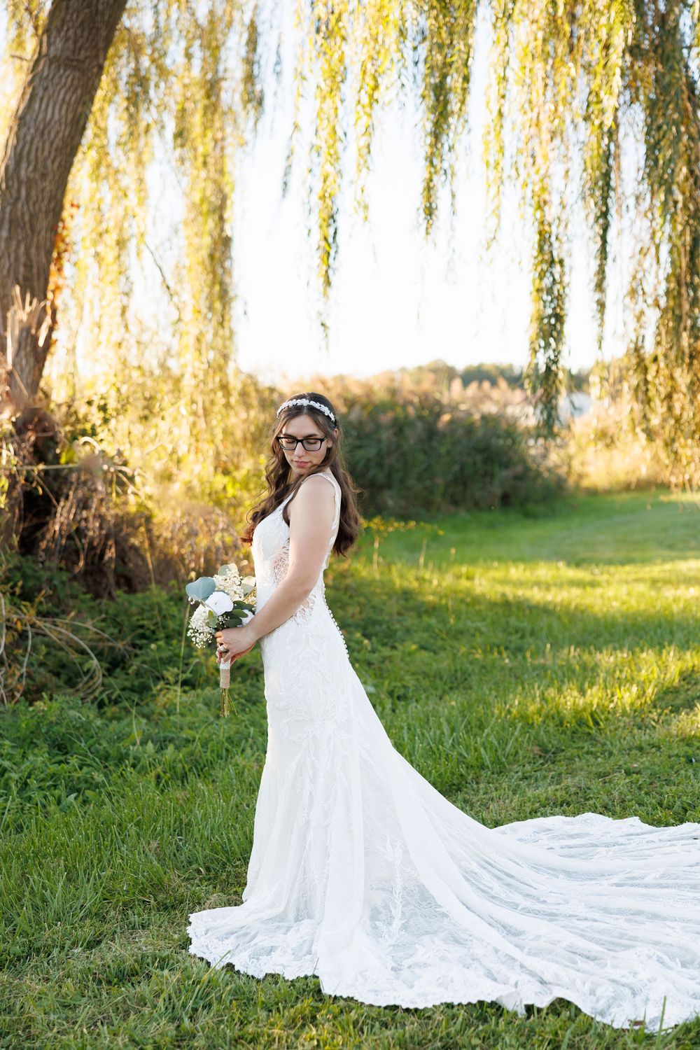 Bride in white lace gown with floral crown holds bouquet under weeping willow tree at golden hour.