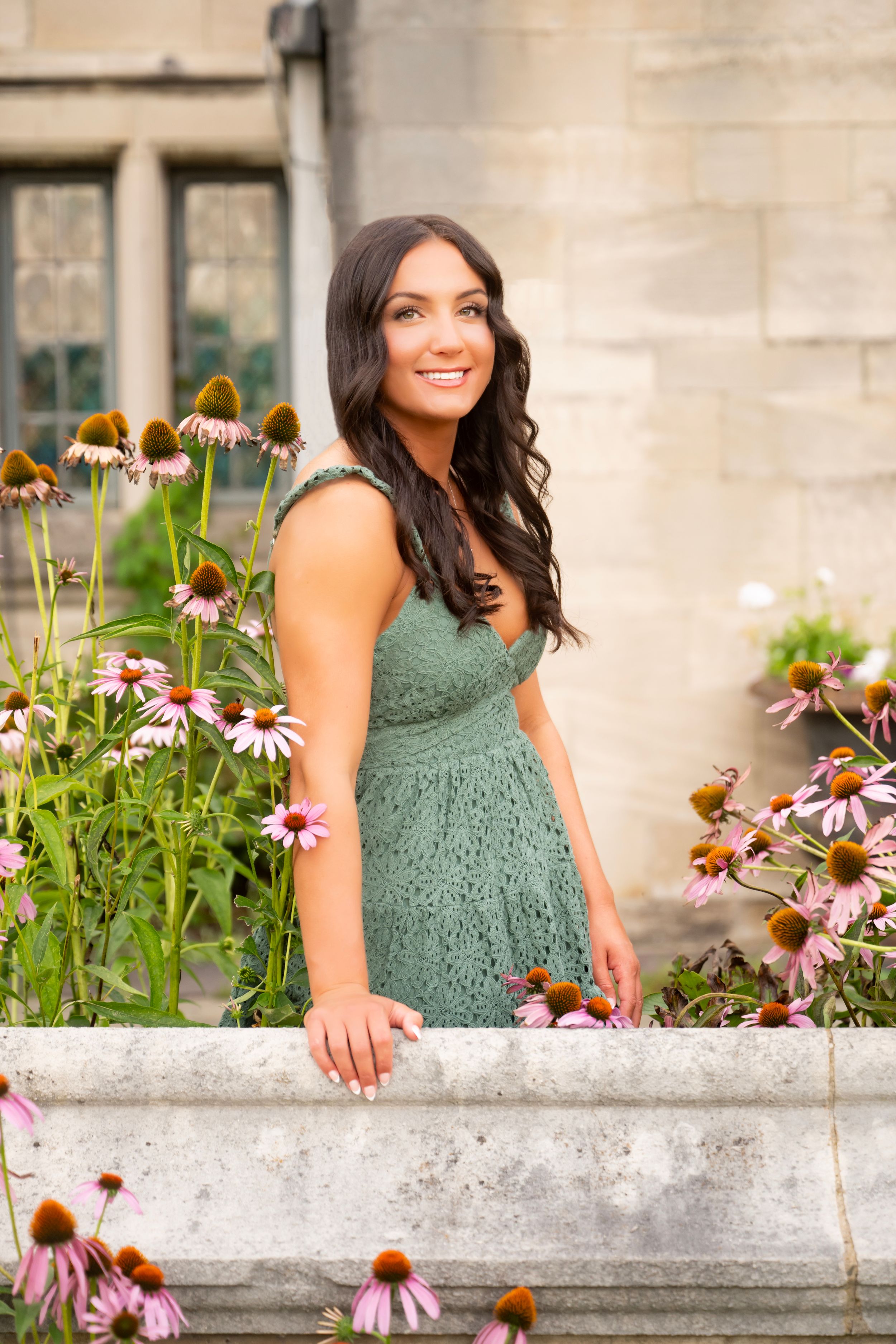 Woman in sage green lace dress poses by stone wall with blooming purple coneflowers in garden setting.