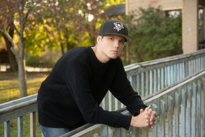 Someone wearing a black sweater and baseball cap leans against a railing in an outdoor setting during autumn.