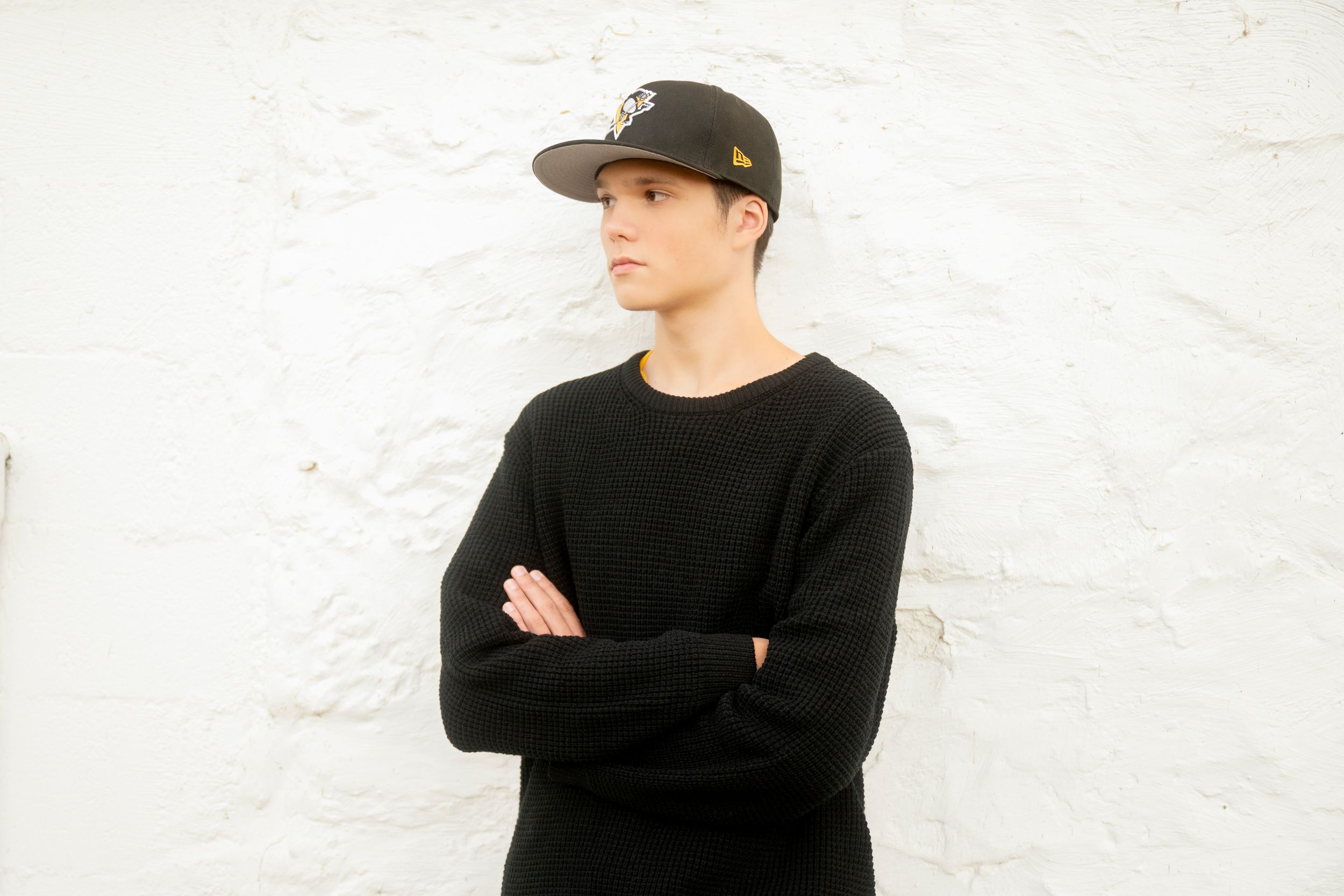 Person wearing a black long sleeve shirt and baseball cap poses against a white textured wall.