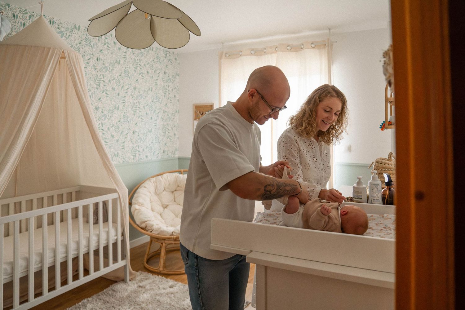Nursery scene with white crib, canopy, and changing table where caretakers tend to an infant in soft natural light.
