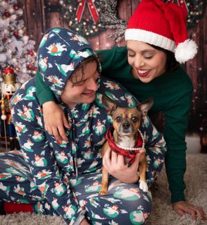 Small dog wearing holiday attire cuddling with owners on festive Christmas backdrop.