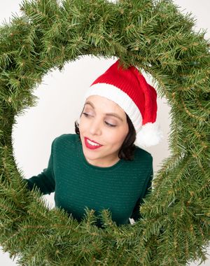 Woman in Santa hat and green sweater posing through a Christmas wreath, smiling with red lips.