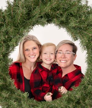 Family of three in matching red plaid shirts smiling through a Christmas wreath holiday photo.