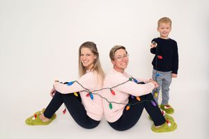 Three people sit back-to-back on the floor wearing matching pink sweaters with string lights wrapped around them.