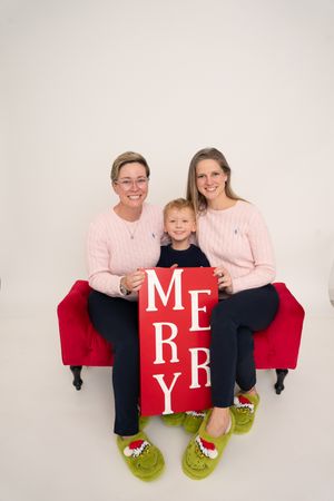 Family of three in matching pink sweaters and Grinch slippers holding a Merry sign on a red couch.