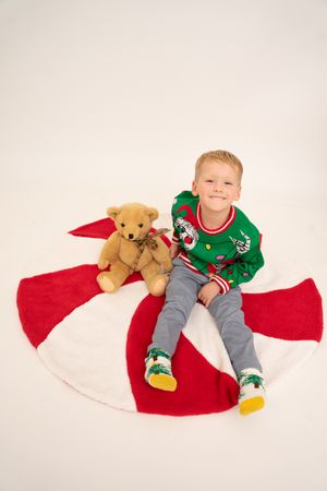 Smiling boy in Christmas sweater sitting on holiday tree skirt with teddy bear, wearing festive socks on white background.