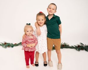 Three siblings wearing holiday outfits pose together against a white background with evergreen branches.