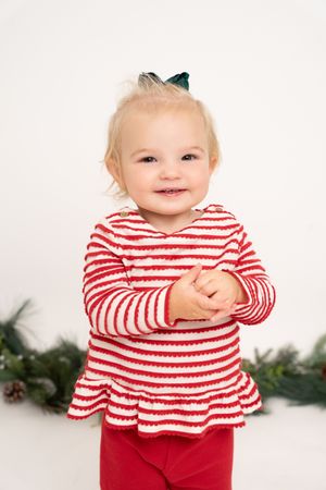 Smiling toddler girl in red striped top and red pants with green bow, posing in front of Christmas greenery.
