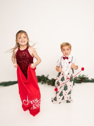 Children in festive holiday outfits pose playfully against a white backdrop with evergreen decor.