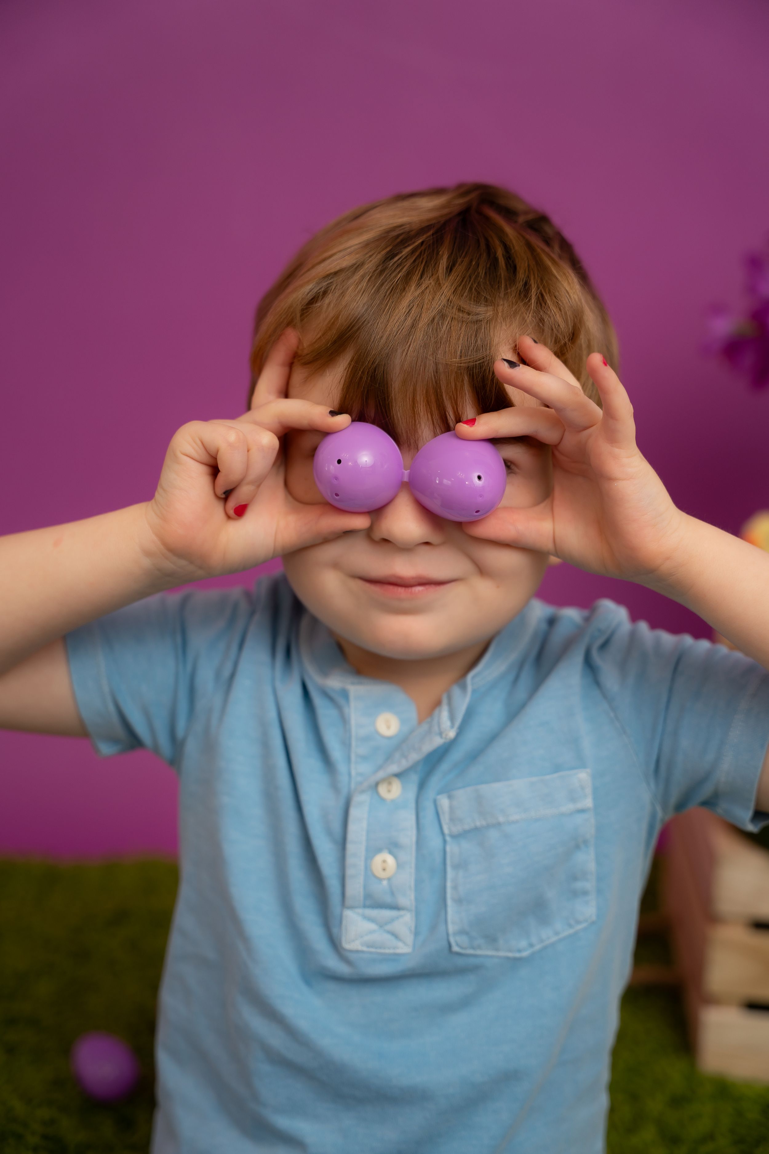 Child wearing blue polo shirt holds purple balls in front of eyes while playing against purple background.