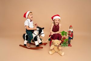 Baby in Santa hat and white outfit sits with teddy bear and Christmas decorations on beige background.