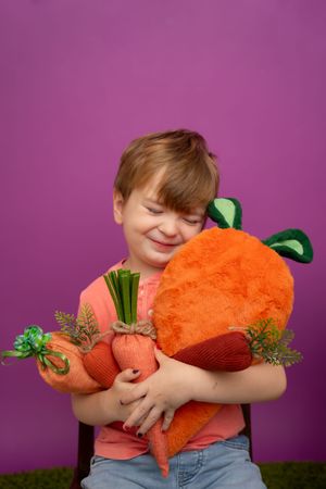Child hugging a large plush orange carrot toy against a vibrant purple background.