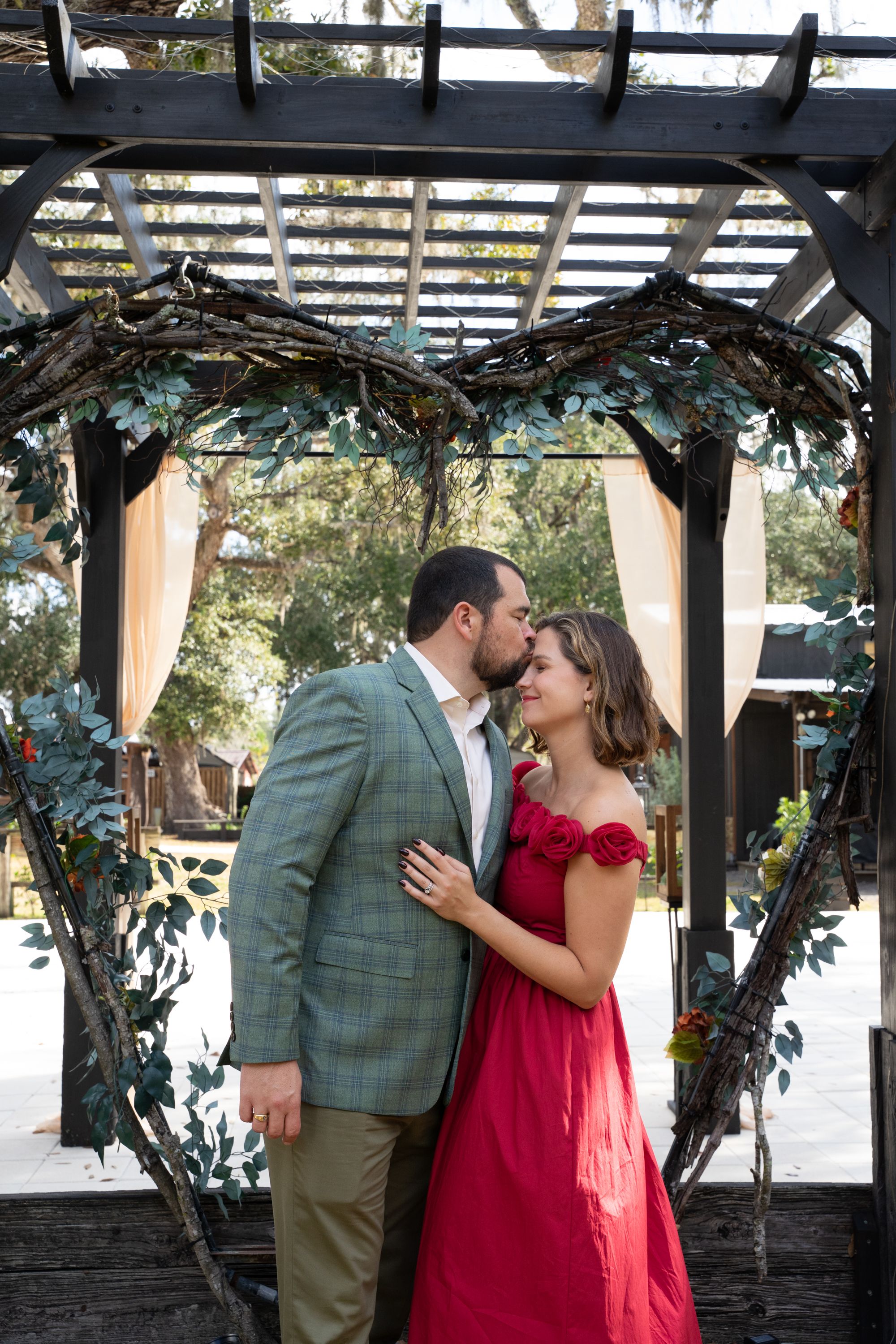 Couple sharing a romantic kiss under a heart-shaped floral arch, woman in red gown, man in gray blazer.