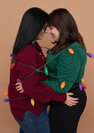 Two people in burgundy and green sweaters share a warm embrace while wrapped in colorful Christmas lights.
