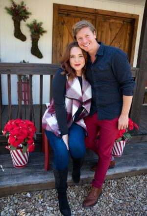 A couple poses on a porch decorated with Christmas stockings and poinsettias for a holiday photo.