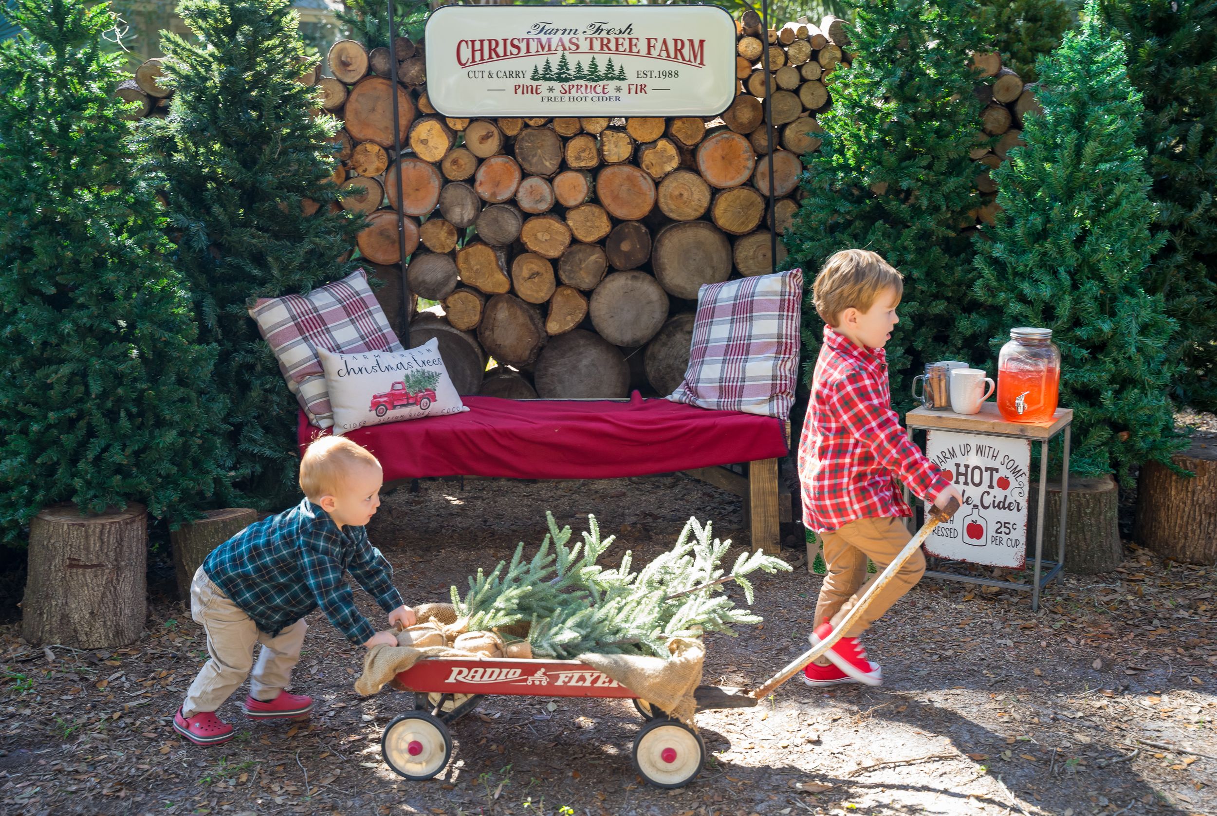 Children pull wagon with Christmas gifts past stacked firewood at holiday event.