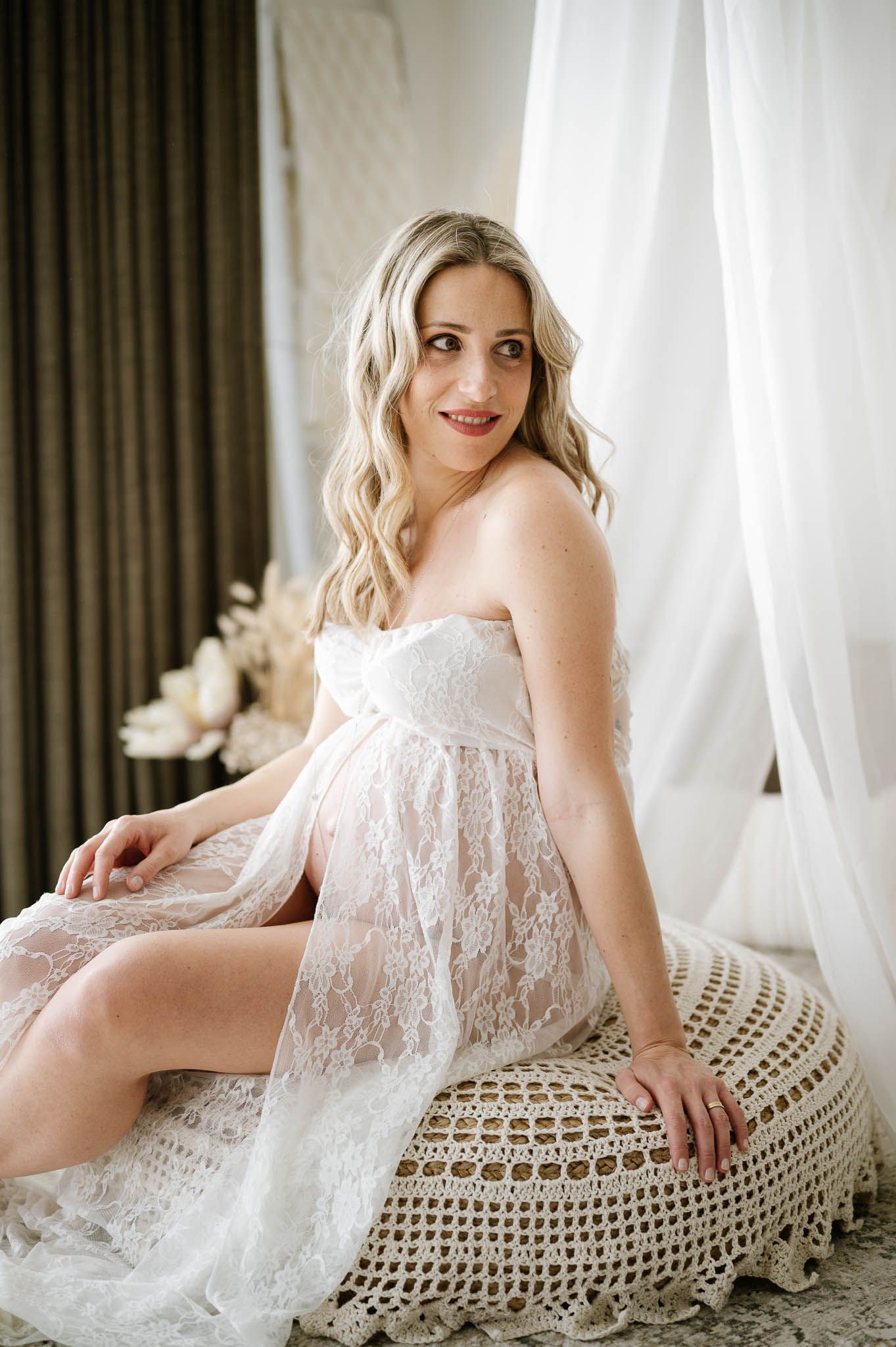 Elegant maternity boudoir photo with soft natural lighting and white sheer curtains.