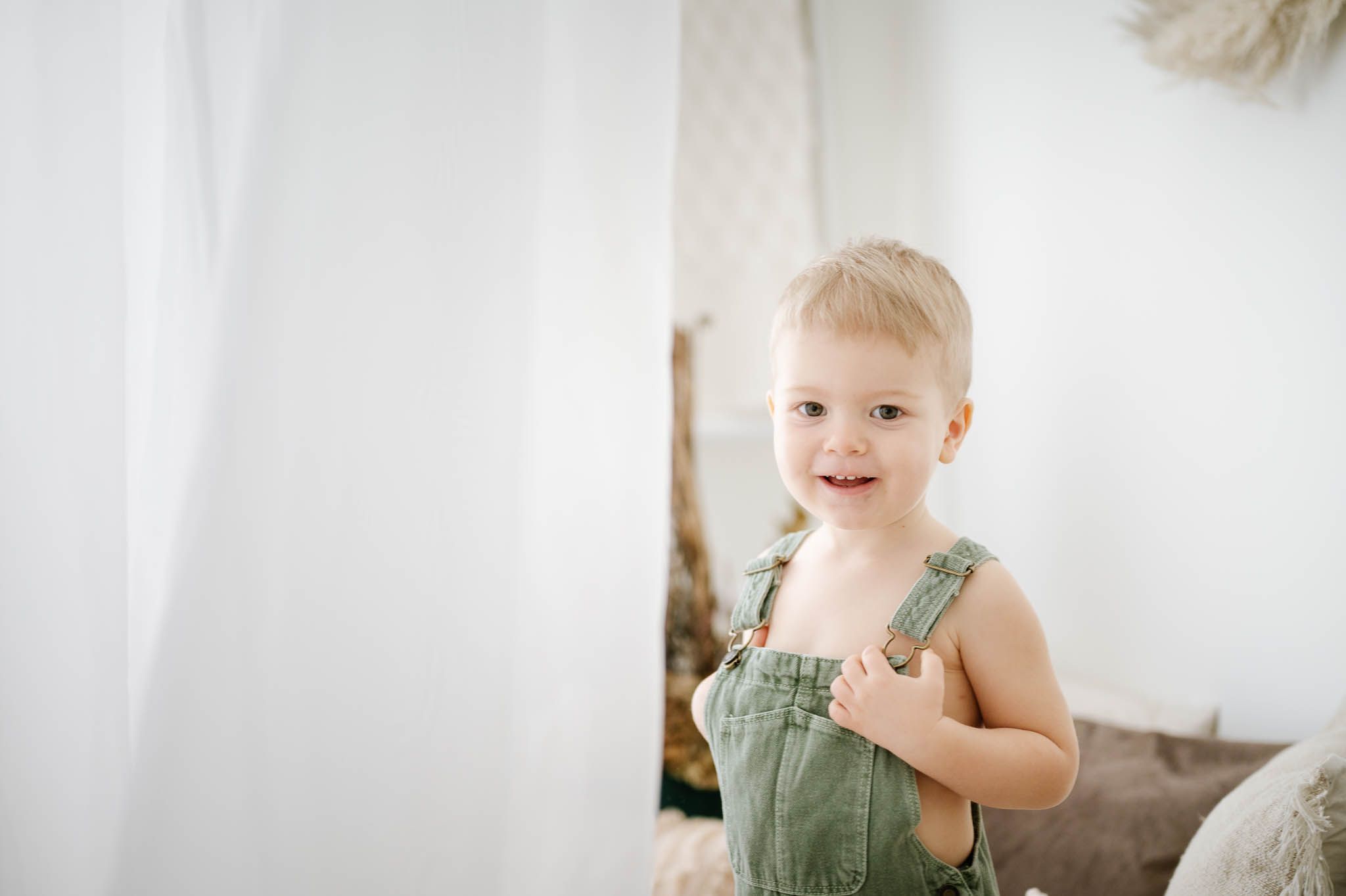 Joyful toddler in green overalls plays peek-a-boo with white curtains.