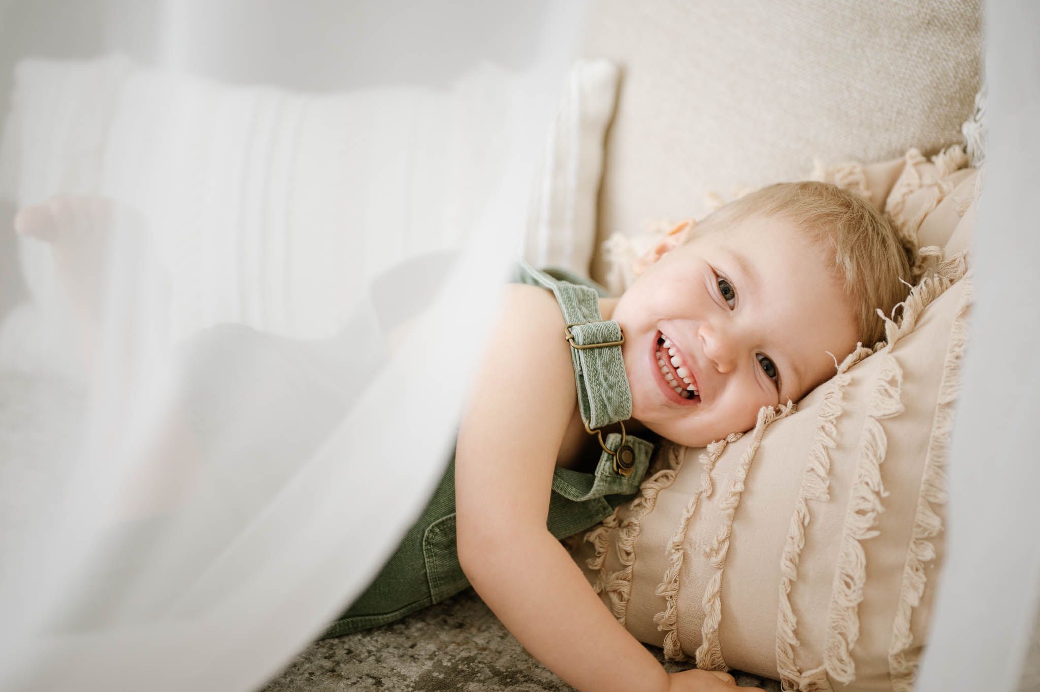 Candid moment of laughing toddler in green outfit laying on cream pillows.