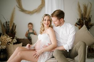 Candid family photo with couple in white and toddler playing in background against light wall decor.