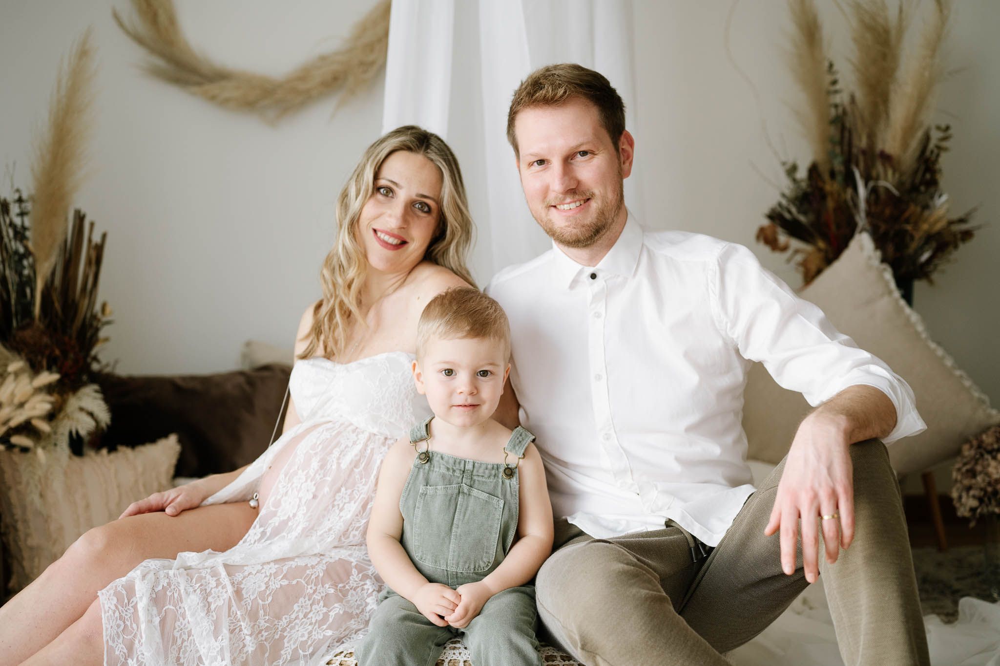 Family portrait of parents in white outfits with toddler in denim overalls against neutral backdrop with dried flowers.