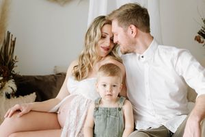 Intimate family moment with parents and toddler in denim overalls on couch with dried floral decor.