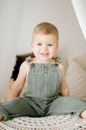 Smiling toddler wearing green denim overalls sitting on white crochet blanket.