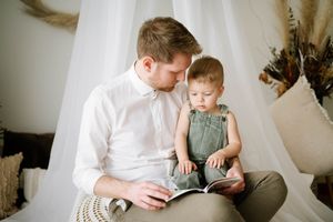 Parent and child reading book together while sitting on textured ottoman near white curtains.