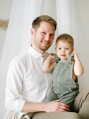 Portrait of parent and toddler in green overalls sitting together against white backdrop.