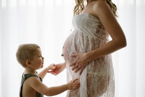 Maternity moment with child touching pregnant belly in front of sheer white curtains.