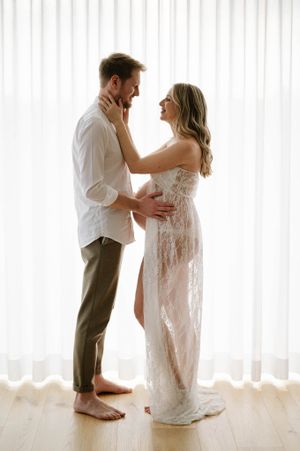 Romantic maternity portrait of couple in white clothing against bright window backdrop.