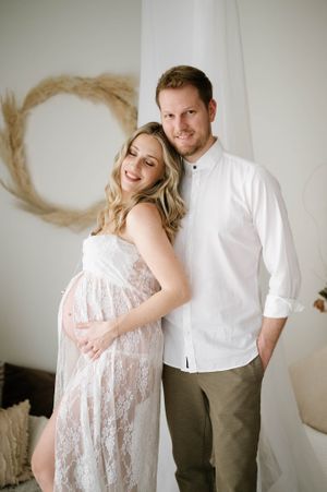 Expectant couple poses together in white and khaki against neutral backdrop with pampas grass decor.