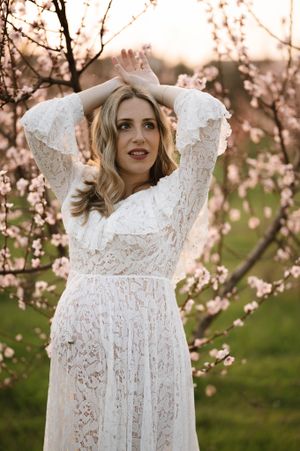 Woman in long sleeve lace dress poses gracefully among blooming cherry trees at sunset.