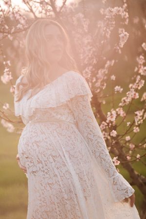 A pregnant woman in a white lace dress poses among blooming cherry blossoms at sunset.