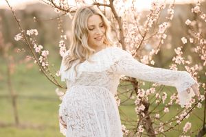 Joyful maternity pose among pink cherry blossoms in flowing white lace gown.