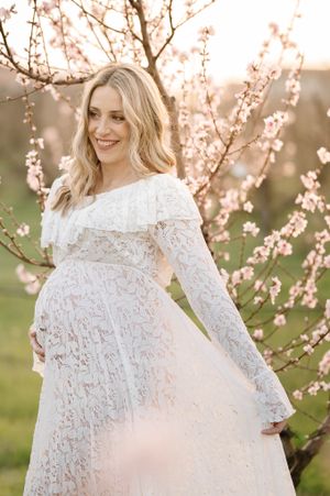 A maternity photo features an ethereal white dress against blooming spring trees at golden hour.