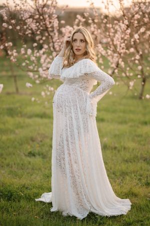 An expectant mother in a flowing white lace gown stands elegantly in a spring orchard.