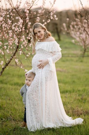 A touching maternity photo with a young child embracing a white-laced dress in a spring orchard.
