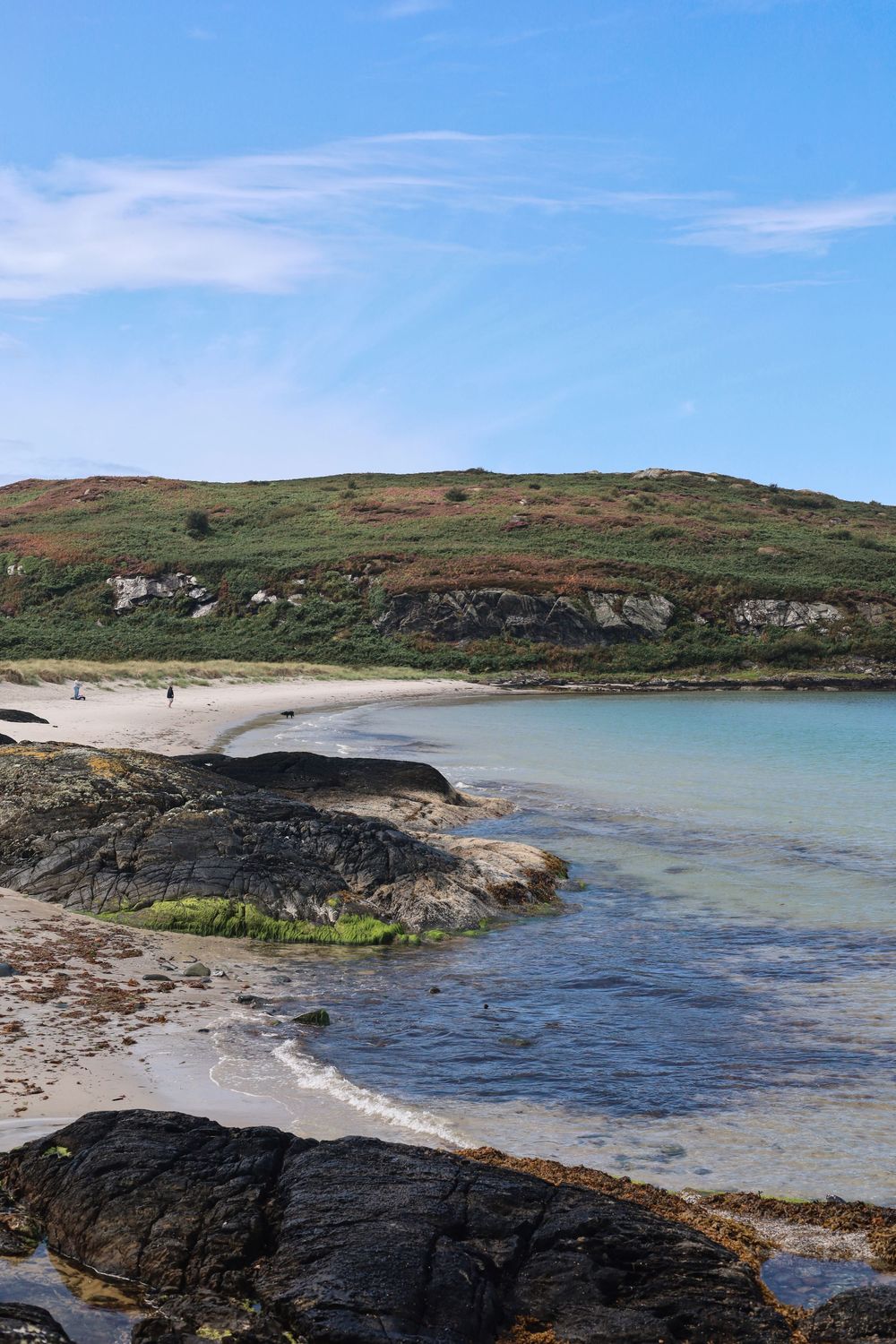 Rugged coastal landscape with a curved sandy beach, rocky outcrops, and turquoise waters under a bright blue sky.