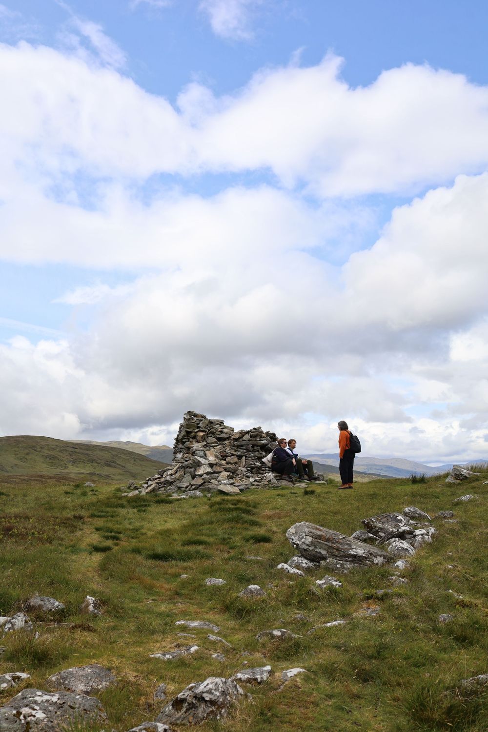 Hikers standing near ancient stone ruins atop a grassy hilltop under cloudy skies in remote countryside.
