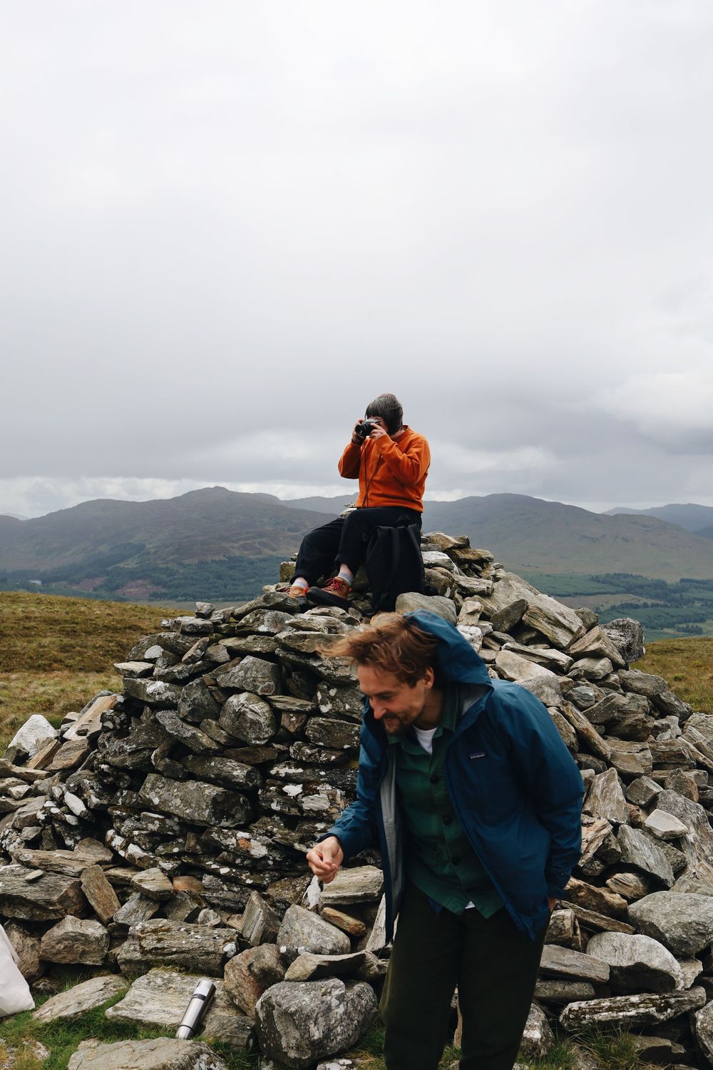 Two hikers examine rock formations at a summit with misty mountains in the background under cloudy skies.